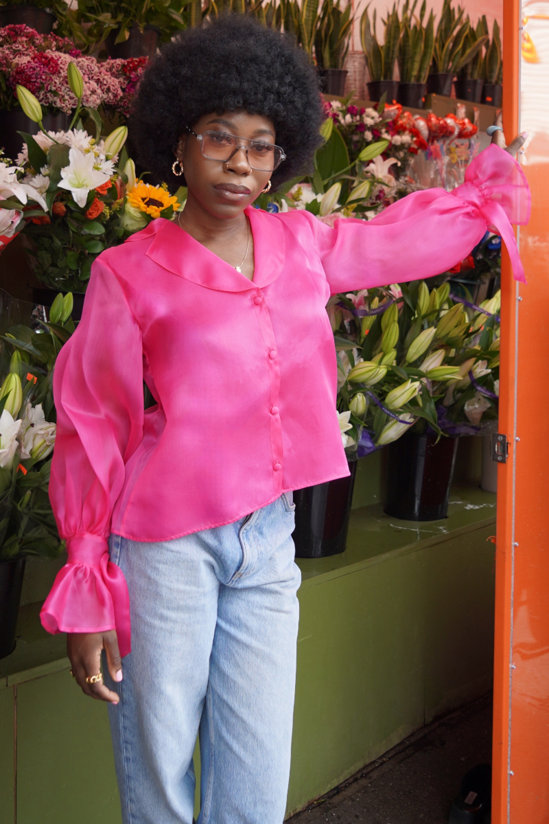 Eine junge modische Afro-Amerikanische Frau, die sich an einem Blumenladen anlehnt und gestylt ist in blauer Jeans und einer Seiden Organza Bluse in Hot Pink von Lola Tong.