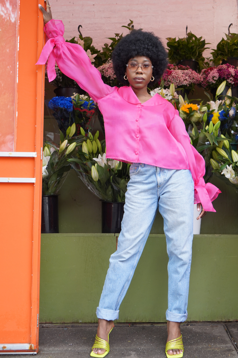 Eine junge modische Afro-Amerikanische Frau, die sich an einem Blumenladen anlehnt und gestylt ist in blauer Jeans und einer Seiden Organza Bluse in Hot Pink von Lola Tong und ihre Haare sind in einem wunderschönen Afro geschnitten.