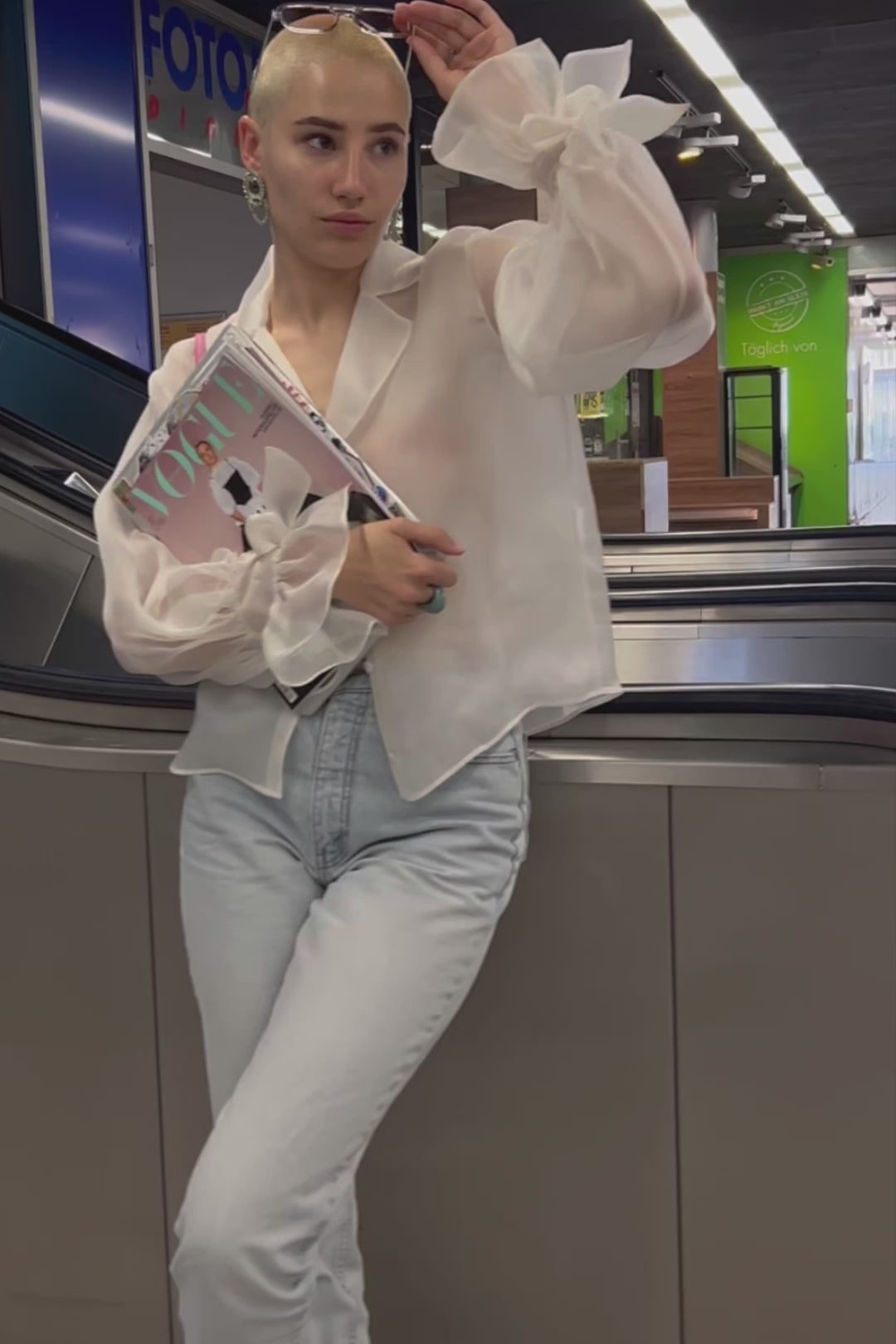 junge Frau mit blonder Kurzhaarfrisur wartet an der Rolltreppe, in ihrer Hand die Deutsche Vogue und gestylt ist sie in blauer Jeans und einer weissen Bluse von Lola Tong.