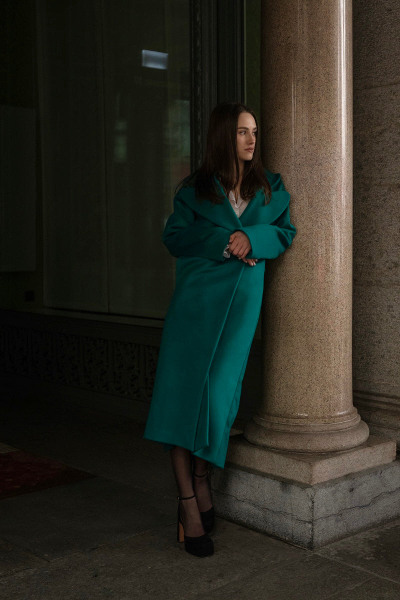 Eine junge Frau in einem grünen langen Mantel von Lola Tong angelehnt an einer Säule in Zürich. 