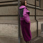 Eine junge elegante Frau mit langen braunen Haaren, angelehnt an einem grauen Gebäude in Zürich. Man sieht die Rückenseite des Mantel in grau, rosa und lila. Sie hat einen teuren Style und trägt einen langen rosa und pinken Mantel von Lola Tong, der aus Kaschmirwolle besteht. 