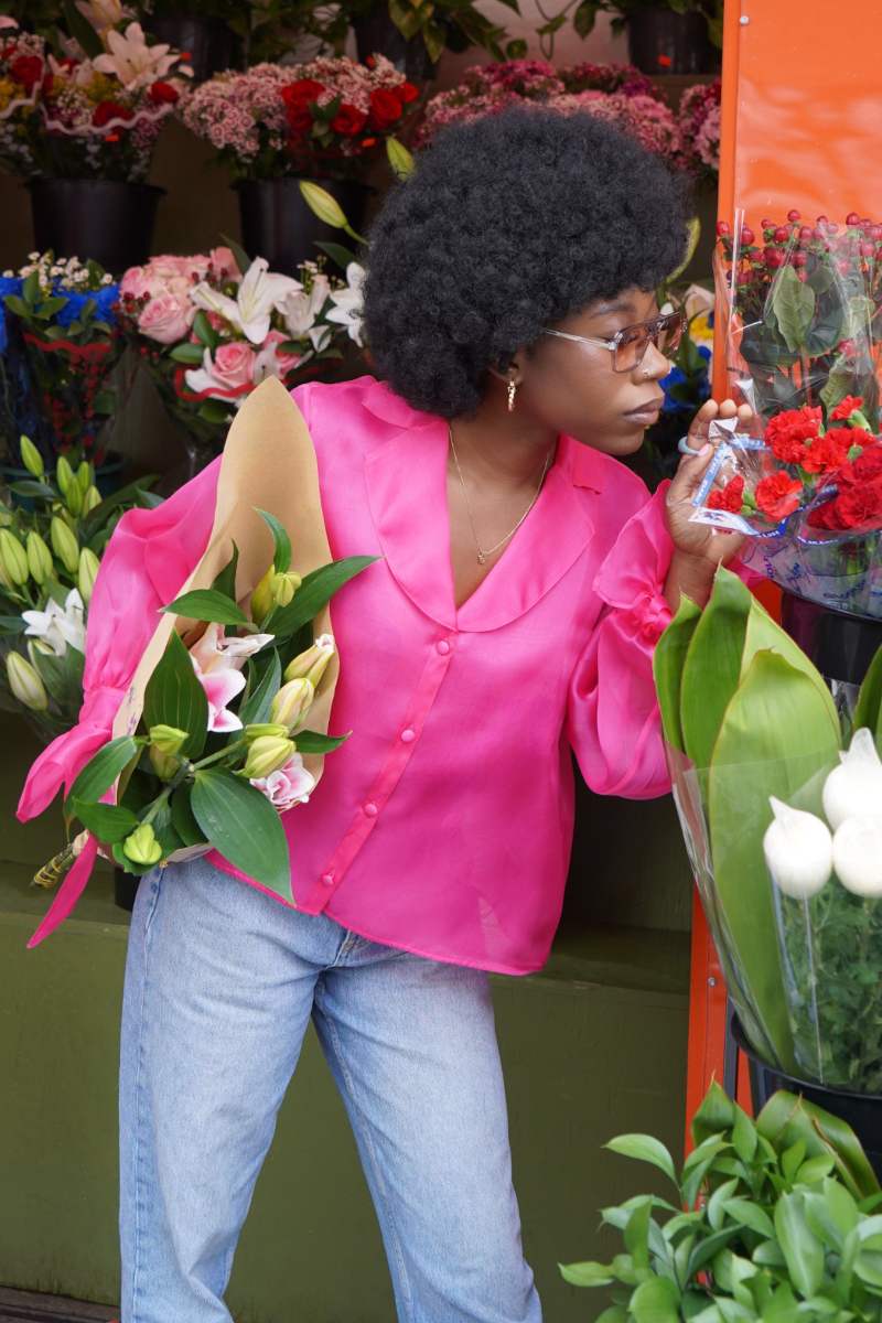 Afro-Amerikanische junge Frau mit einem Blumenstrauss in der Hand, während Sie eine blaue Jeans an hat und eine Seiden Organza Bluse in Hot Pink von Lola Tong.