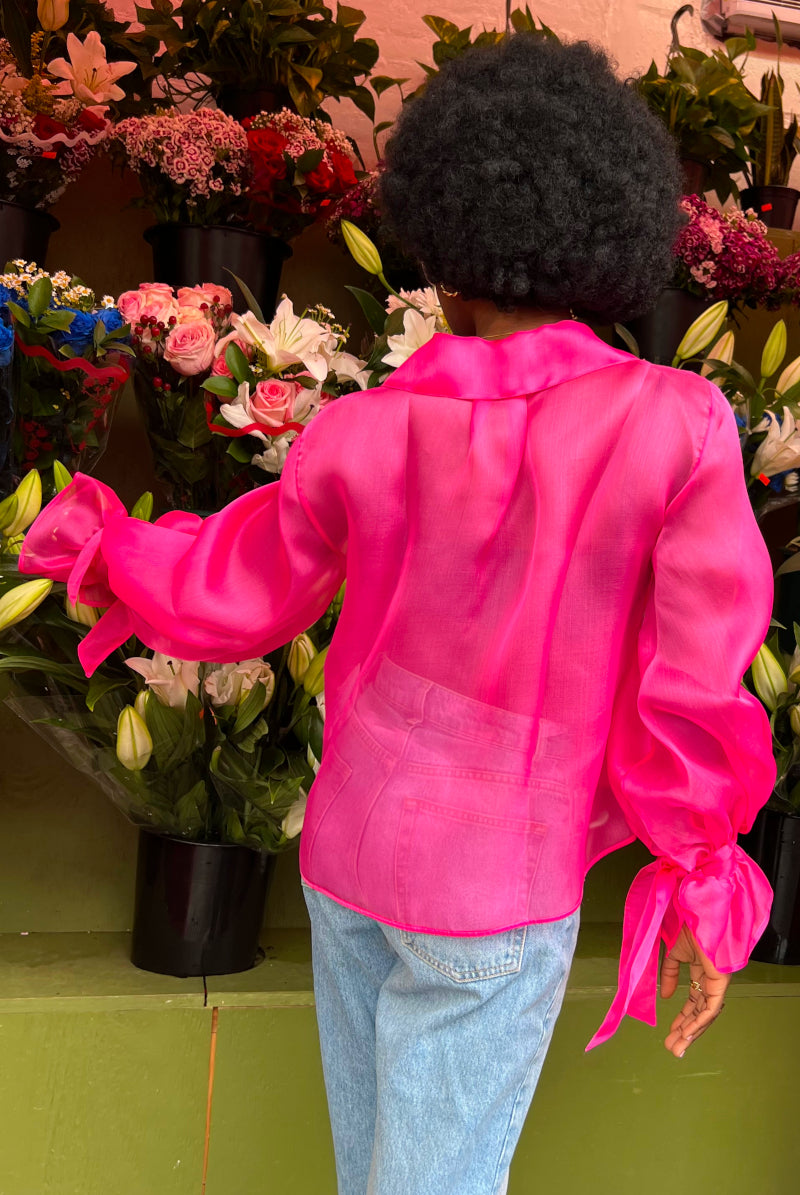 Rückenansicht einer Afro-Amerikanischen junge Frau in einem Blumenladen, während sie eine blaue Jeans an hat und eine Seiden Organza Bluse in Hot Pink von Lola Tong.