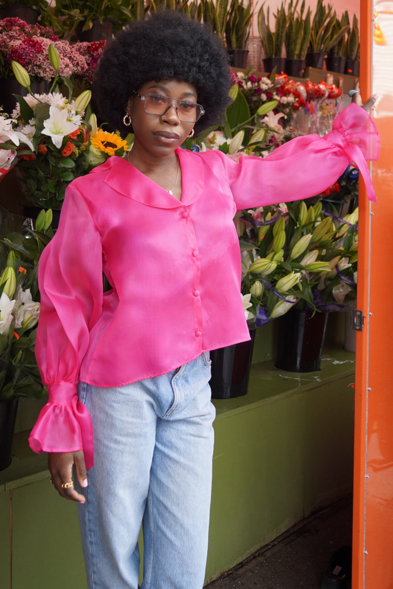 Eine junge modische Afro-Amerikanische Frau, die sich an einem Blumenladen anlehnt und gestylt ist in blauer Jeans und einer Seiden Organza Bluse in Hot Pink von Lola Tong. 