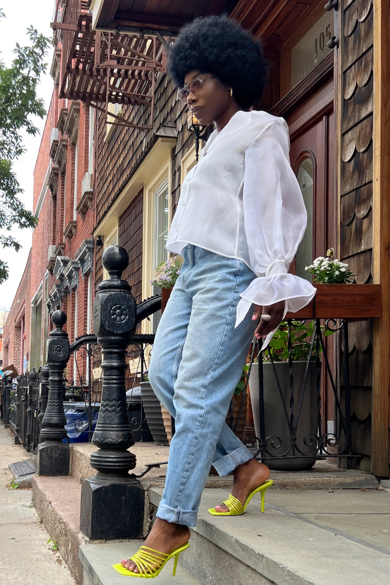 Eine junge Amerikanerin verlässt ihre Wohnung in einer blauen Jeans, der weißen Lola Tong-Bluse aus Seidenorganza und lindgrünen Absätzen. 
