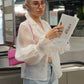 Junge Frau liest die Deutsche Vogue während sie am Rollband auf jemanden wartet. Dabei hat die eine durchsichtige weiße Bluse von Lola Tong an, eine blaue Jeans, eine rosa Handtasche, eine Sonnenbrille und auffallende Ohrringe.