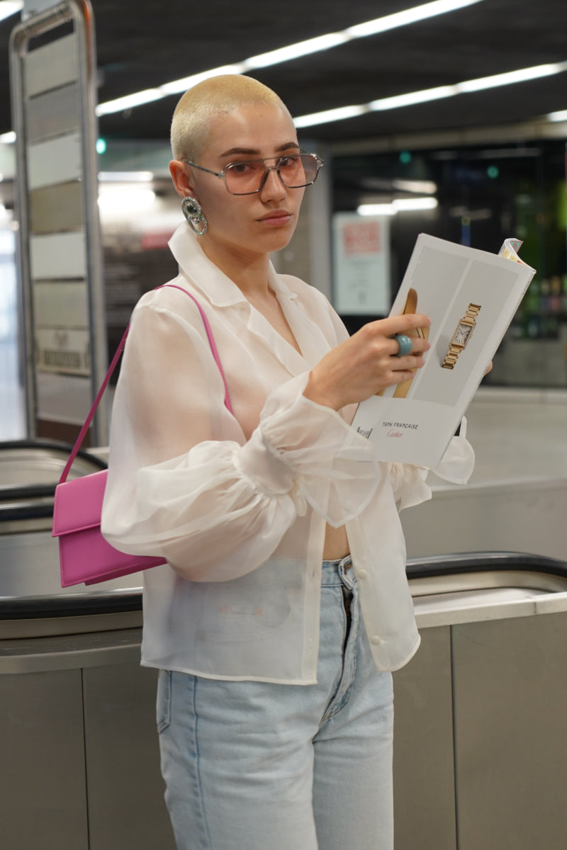 Junge Frau liest die Deutsche Vogue während sie am Rollband auf jemand wartet. Dabei hat die eine durchsichtige weisse Bluse von Lola Tong an, eine blaue Jeans, eine rosa Handtasche, eine Sonnenbrille und auffallende Ohrringe.